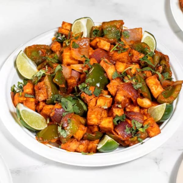 Indian paneer dish with bell peppers, onions, cilantro, and lime wedges on a white plate.